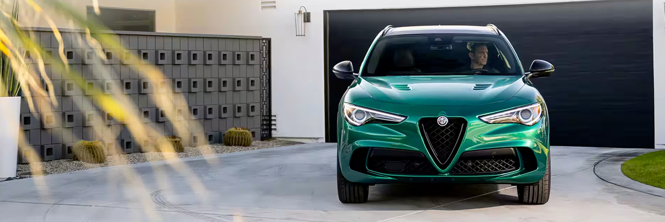 A green 2023 Alfa Romeo Stelvio Quadrifoglio parked in the driveway of a contemporary home with a driver inside.
