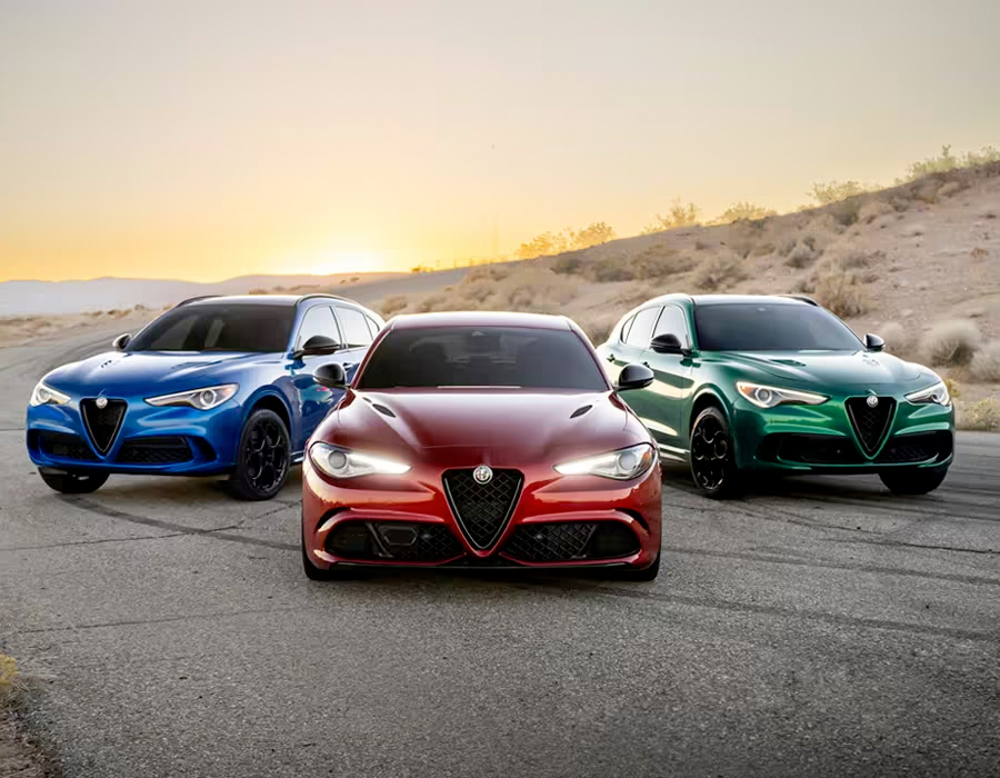 Three 2023 Alfa Romeo models parked beside a hill with the sun rising in the background. From left to right: a blue 2023 Alfa Romeo Stelvio Quadrifoglio, a red 2023 Alfa Romeo Giulia Quadrifoglio and a green 2023 Alfa Romeo Stelvio Quadrifoglio.