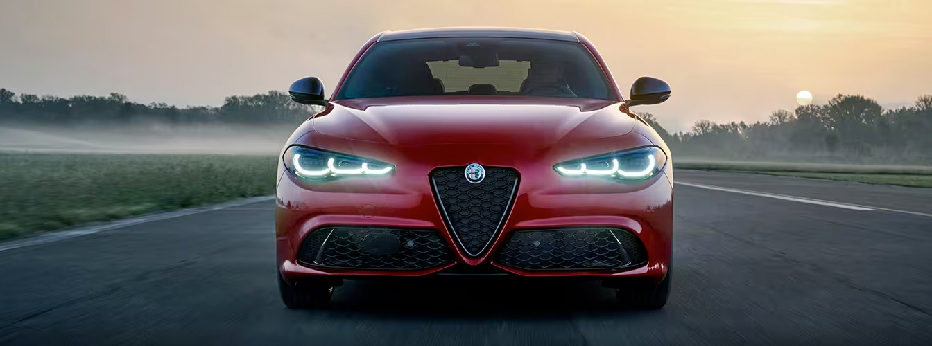 The front end of a red 2024 Alfa Romeo Giulia Veloce being driven on a track at sunset.
